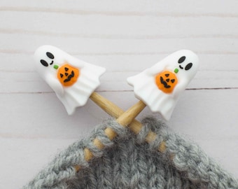 May include: Two white ghost-shaped stitch markers with orange pumpkin details are crossed over a gray knitted fabric. The markers are on wooden knitting needles. The background is a light wood surface. Halloween-themed knitting accessories.