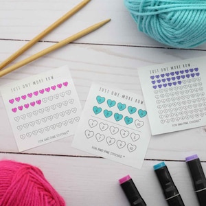 May include: Three square white paper trackers with the text JUST ONE MORE ROW and FOX AND PINE STITCHES. Each tracker features rows of heart outlines with numbers inside. Some hearts are colored in pink, teal, or purple ink.