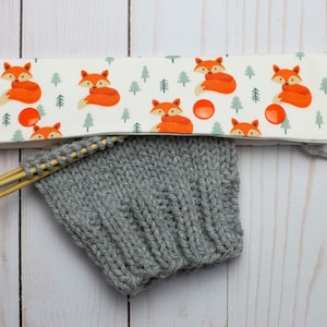 May include: A white fabric needle minder with a repeating pattern of orange foxes and green trees. The minder has three orange buttons. The minder is sitting on a wooden surface with a gray knitted piece and wooden knitting needles.