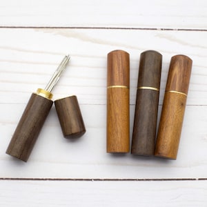 May include: Four cylindrical wooden needle cases with brass accents. One case is open, revealing a silver metal needle inside, while the other three cases are closed. The wood features varying shades of brown with visible natural grain patterns.