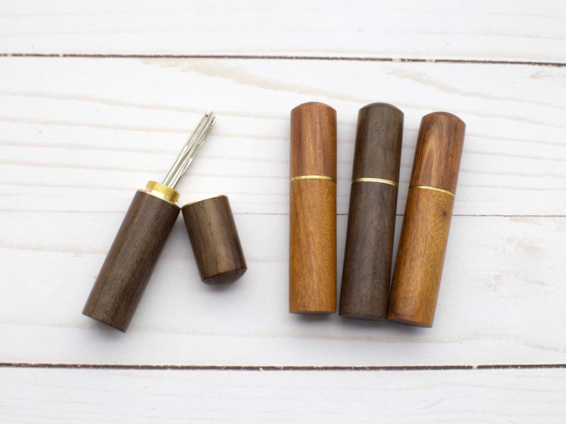 May include: Four cylindrical wooden needle cases with brass accents. One case is open, revealing a silver metal needle inside, while the other three cases are closed. The wood features varying shades of brown with visible natural grain patterns.