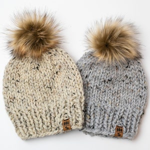 May include: Two knitted hats with faux fur pom poms. The hats are beige and gray with a brown leather patch on the side that says "Pine & Oak".