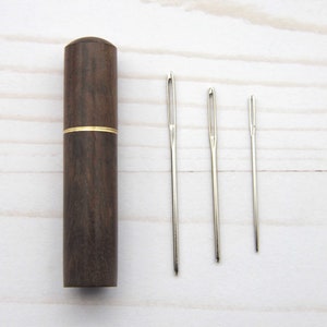 May include: A dark brown cylindrical wooden needle case with a thin gold-colored metal band around the center, accompanied by three silver-colored metal sewing needles of varying lengths arranged in a row to the right of the case.