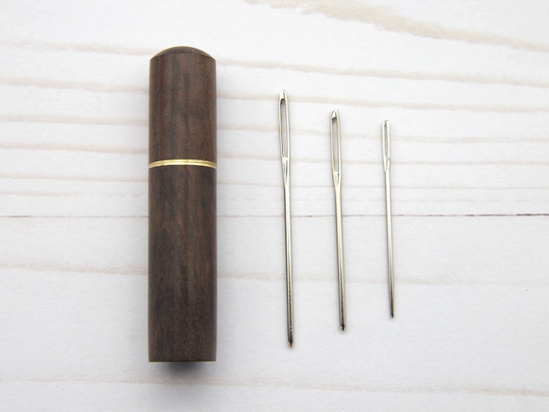 May include: A dark brown cylindrical wooden needle case with a thin gold-colored metal band around the center, accompanied by three silver-colored metal sewing needles of varying lengths arranged in a row to the right of the case.