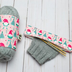 May include: A gray yarn ball wrapped with a pink fabric featuring gnomes and Christmas trees. A matching needle case with wooden needles and a knitted gray cuff are also present. The fabric has a small tag.