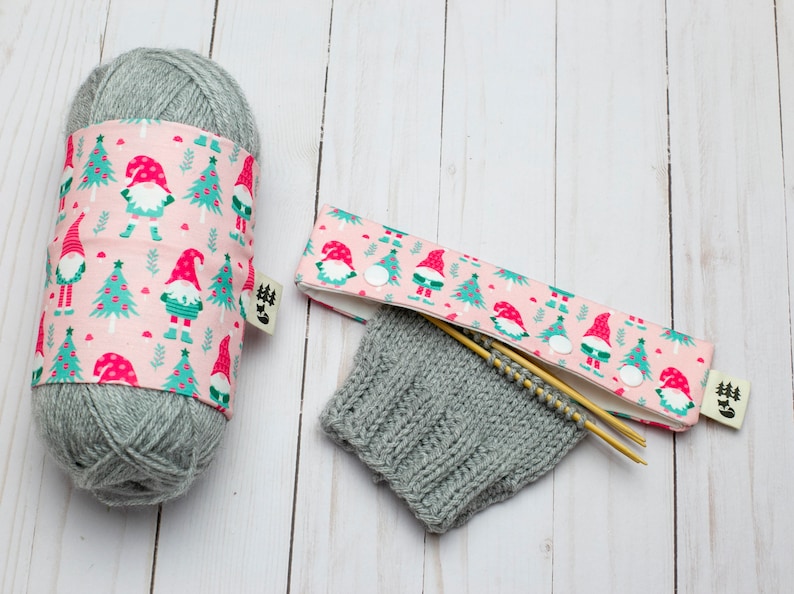 May include: A gray yarn ball wrapped with a pink fabric featuring gnomes and Christmas trees. A matching needle case with wooden needles and a knitted gray cuff are also present. The fabric has a small tag.