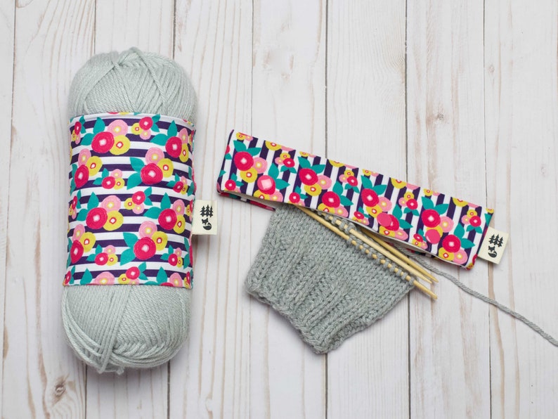 May include: A skein of light blue yarn with a floral patterned fabric cover, a knitted hat, and two wooden knitting needles.
