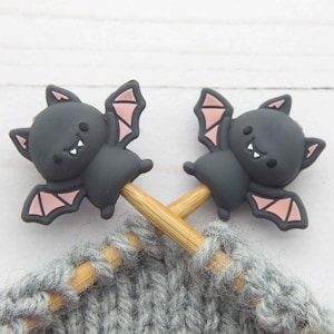 May include: Two dark gray bat-shaped stitch markers with pink wing details are attached to wooden knitting needles, resting on a gray knitted fabric. The bats have smiling faces. The background is a white wooden surface.