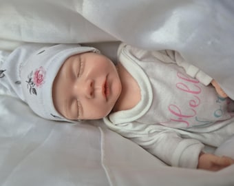 May include: A realistic baby doll wearing a white onesie with the word "Hello" in pink script. The doll is also wearing a white hat with a pink rose design. The baby is nestled under a white blanket.