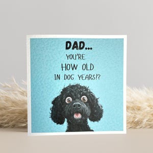 May include: A square greeting card with a light blue background and the text "DAD... YOU'RE HOW OLD IN DOG YEARS!?" A black dog with wide eyes and an open mouth is at the bottom of the card. The card is a humorous gift.