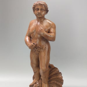 May include: A wooden sculpture of a nude male figure standing on a seashell. The figure is carved in a realistic style and has a youthful appearance.