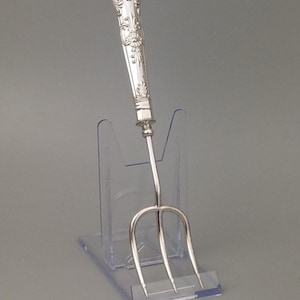 May include: A silver-toned serving fork featuring an ornate, embossed handle with intricate scrollwork and shell motifs. The utensil has three long, slender tines and a polished metallic finish, displayed upright on a clear plastic stand.