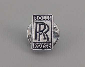 May include: A silver Rolls Royce lapel pin. The pin displays the Rolls Royce logo, with the words "ROLLS" above and "ROYCE" below. The logo is a stylized "R" inside a rectangular frame, a classic emblem.