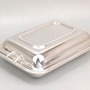 Op de afbeelding: Een rechthoekige zilverkleurige serveerschaal met bijpassend deksel. Het stuk heeft een gepolijste metalen afwerking en een decoratieve kralenrand langs de randen van zowel het deksel als de basis. Aan de zijkanten zijn twee kleine metalen handgrepen bevestigd.