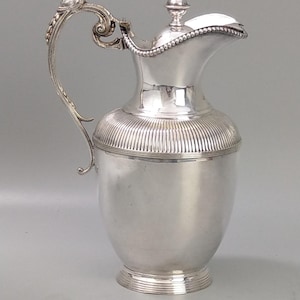 Puede incluir: Una jarra plateada con un asa decorativa y una tapa. La jarra tiene un cuerpo bulboso con detalles acanalados verticales cerca de la parte superior. El asa es ornamentada y la tapa está rematada con un pequeño adorno.