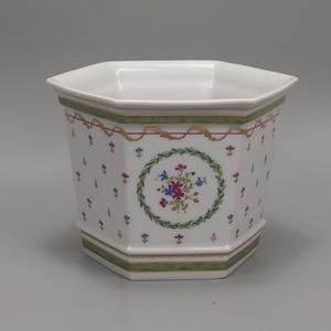 Op de afbeelding: Witte, achthoekige keramische plantenbak met bloemen- en lintontwerp. De plantenbak heeft een groene en gouden rand, met een centrale bloemenkrans. Kleine blauwe, rode en gele bloemen zijn verspreid.