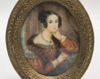 May include: An oval portrait miniature painting of a person with dark hair, wearing a reddish-brown dress with a fur collar and a multi-colored gemstone necklace. The portrait is encased in an ornate, metallic gold-toned frame with a Greek key border.