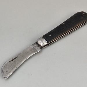 May include: A vintage folding pocket knife with a black wooden handle and a curved blade. The blade is marked with the text "G.R.O."