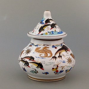 Può includere: Un vaso di ceramica bianca con un coperchio, decorato con un motivo colorato di vita marina. Il vaso presenta cavallucci marini, pesci e stelle. Il coperchio è a forma di cono ed è anche decorato con vita marina.