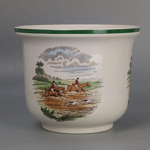 Puede incluir: Un macetero de cerámica blanca con un borde verde presenta una escena de caza con jinetes a caballo y perros. La escena se desarrolla sobre un fondo de cielo nublado y colinas.