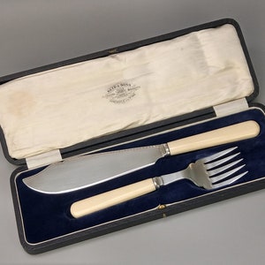 Puede incluir: Un estuche de terciopelo negro forrado con satén blanco. El estuche contiene un cuchillo y un tenedor de plata con mangos de marfil. El estuche tiene una etiqueta que dice "Reid & Sons, Sheffield, England".