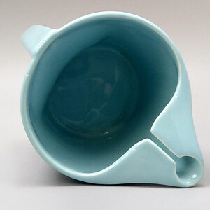 May include: A light blue ceramic pitcher with a unique spout design. The pitcher has a handle and a rounded body. The interior is a darker shade of blue. The pitcher is likely used for pouring liquids.