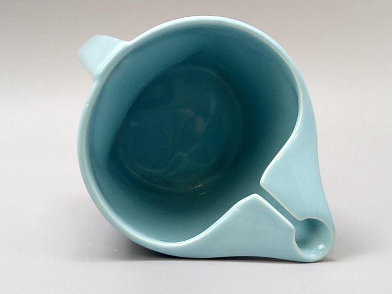 May include: A light blue ceramic pitcher with a unique spout design. The pitcher has a handle and a rounded body. The interior is a darker shade of blue. The pitcher is likely used for pouring liquids.