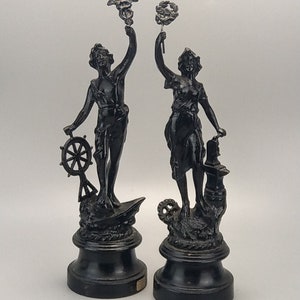 May include: Two black statues of classical figures. One figure holds a ship's wheel and stands on a boat. The other figure holds a wreath and stands near a gear. Both figures are on a round base.