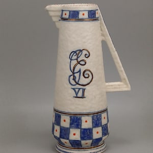 Puede incluir: Jarra de cerámica blanca con detalles azules y marrones. La jarra presenta un patrón de cuadros azules y blancos en la base y un asa decorativa. Un monograma azul y marrón y el número romano VI están en el frente.