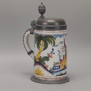 Könnte beinhalten: Ein Vintage-Bierkrug aus Keramik mit einem Klappdeckel aus Zinn. Der Krug zeigt eine handgemalte Szene einer tropischen Landschaft mit Palmen und einer Kirche. Der Griff ist weiß mit einem dekorativen Muster.