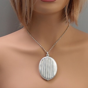 May include: Silver-toned oval locket pendant necklace. The locket has a vertically striped design on the front. The necklace chain is a delicate, linked style. The pendant hangs from the chain, resting on a person's chest. Jewelry.