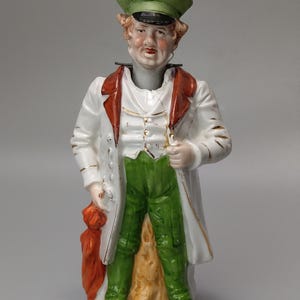 May include: A porcelain figurine of a man wearing a white coat, green trousers, and a green hat. He is holding a red bag in his left hand and has a gold trim on his coat.