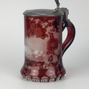 May include: A ruby-red glass beer stein with a silver metal lid and handle. The stein features an etched design of a building and floral patterns. The base has a decorative, scalloped edge. A classic piece for beer enthusiasts.