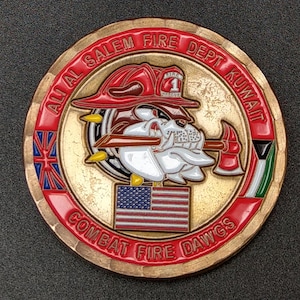 May include: A circular coin in bronze with red and gold detailing. It features a bulldog wearing a firefighter's helmet and holding an axe. The text "All Al Salem Fire Dept Kuwait" and "Combat Fire Dawgs" are visible. The American flag is also displayed.