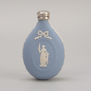 Op de afbeelding: Een lichtblauwe fles met een zilveren dop en witte reliëfdetails. De fles heeft een centraal ovaal met een klassieke figuur en een strik aan de bovenkant. De randen zijn ook versierd met witte reliëfs.