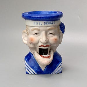 May include: A ceramic ashtray shaped like a sailor's head with a blue and white sailor hat. The ashtray has the words "H.M.S. BREEZY" written on the hat.