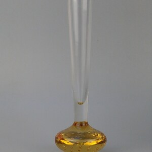 Könnte beinhalten: Eine klare Glas-Blumenvase mit einem runden, bernsteinfarbenen Sockel. Der Sockel ist mit kleinen Luftblasen strukturiert.