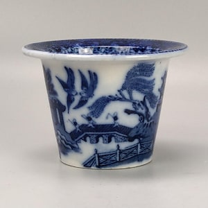 May include: A small, white ceramic planter with a blue and white floral design. The planter is decorated with a traditional Chinese landscape scene featuring a bridge, trees, and birds.