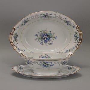 May include: A white porcelain serving dish set with a floral design in blue and purple. The set includes an oval serving platter with gold trim and handles, a gravy boat, and a saucer. The floral pattern is repeated on each piece.