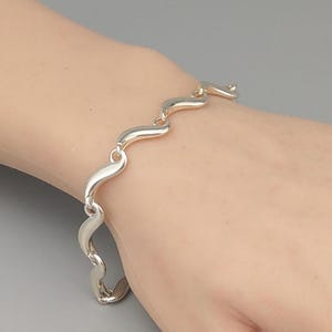 May include: A silver bracelet with a wavy design. The bracelet is composed of interconnected, curved links that create a flowing, organic pattern. The links are smooth and polished, reflecting light. The bracelet is worn on a wrist.