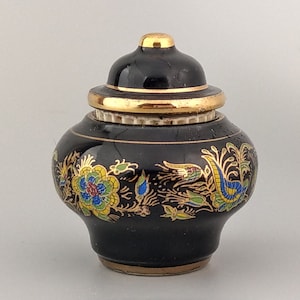 Può includere: Un piccolo vaso in ceramica nera con coperchio, ornato con finiture dorate e disegni floreali e di uccelli colorati. Il vaso ha un corpo arrotondato e un piccolo coperchio dorato. I dettagli includono blu, giallo e verde.