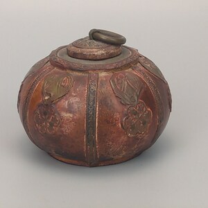 May include: A round, brown leather box with intricate embossed designs and a metal ring handle on the lid.