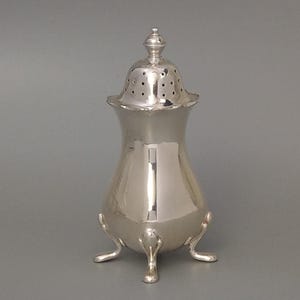 Könnte beinhalten: Ein silberfarbener, verzierter Shaker mit perforiertem Deckel und dekorativem Knauf. Der Shaker hat einen bauchigen Körper, der von drei kleinen Füßen getragen wird. Das Design ist elegant und klassisch.