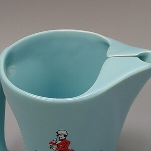 May include: A light blue pitcher with a curved handle and a spout. The pitcher has a wide opening and a decorative image of a person in a red coat. The pitcher is made of a smooth, glossy material.