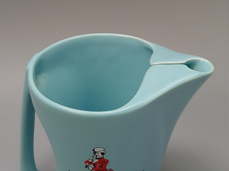 May include: A light blue pitcher with a curved handle and a spout. The pitcher has a wide opening and a decorative image of a person in a red coat. The pitcher is made of a smooth, glossy material.
