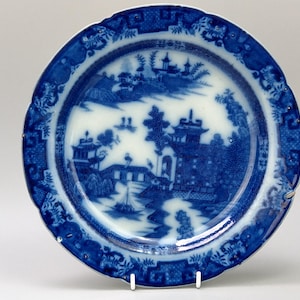 May include: A blue and white porcelain plate with a detailed landscape scene. The plate features a scalloped edge and a central design of pagodas, trees, and a bridge. The plate is supported by a white stand.
