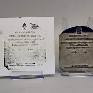 May include: A white certificate of authenticity for a Royal Crown Derby Heraldic Derbyshire Stag paperweight, limited edition of 950, alongside a matching metal printing plate with the same text and logo, both displayed on clear plastic stands.