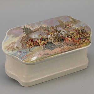 Puede incluir: Caja de baratijas rectangular de cerámica blanquecina con tapa decorativa. La tapa presenta una escena detallada de una batalla con figuras a caballo. La caja tiene una base curva y un acabado ligeramente iridiscente.