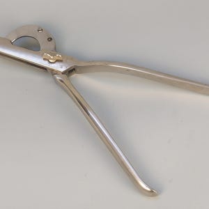 May include: A silver surgical tool with a curved handle and a circular cutting blade. The tool has a small lever on the handle.