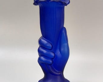 May include: A cobalt blue glass vase in the shape of a hand holding a tulip-shaped vessel. The vase has a ruffled top and a detailed base. The hand is sculpted with visible fingers and a textured surface. The vase is approximately 7 inches tall.
